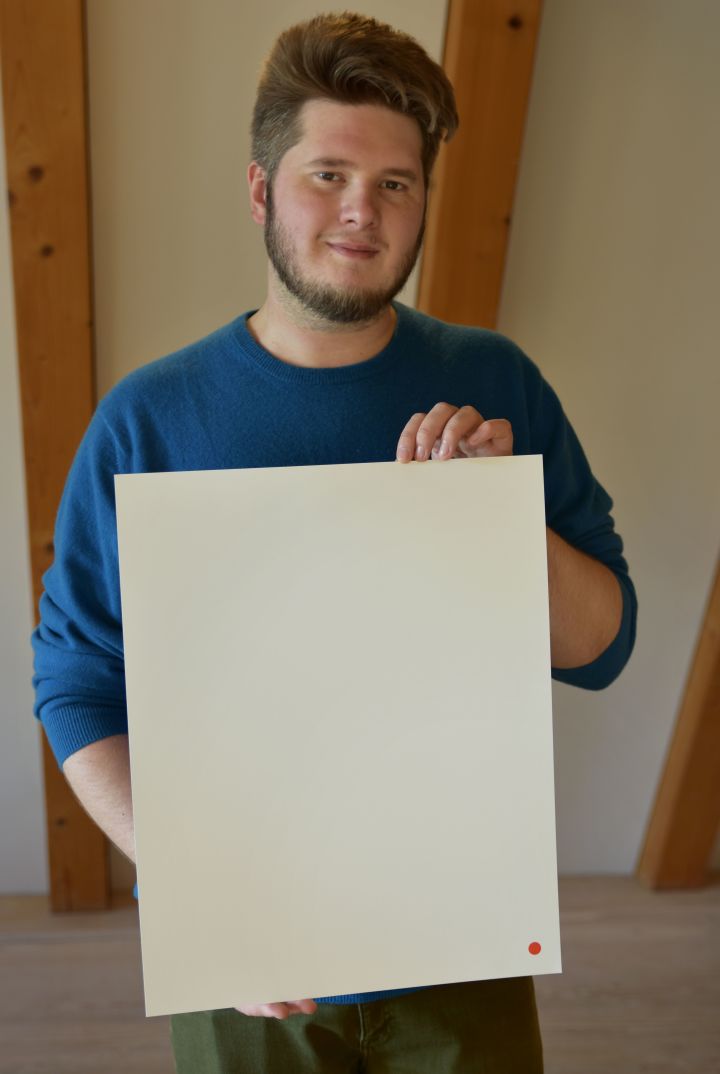 Künstler mit seiner Schenkung in der Hand, junger Mann in blauem Pullover, roter Punkt auf weißem Blatt